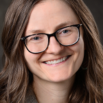 A white woman with long brown hair and dark-rimmed glasses smiles