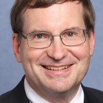 A white man wearing a suit and tie wearing thin-rimmed rectangular glasses with short brown hair smiles