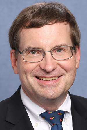 A white man wearing a suit and tie wearing thin-rimmed rectangular glasses with short brown hair smiles